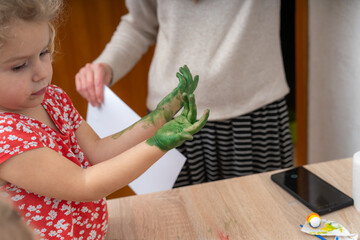Child enjoys art activity with green paint on hands at home in the afternoon with an adult nearby...