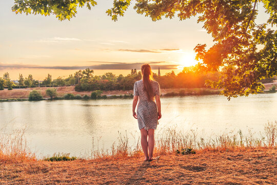Young woman from behind looking at sunrise over calm water, surrounded by soft golden light. Concept of freedom, new chapter, mindfulness and connection with nature