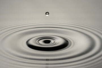 A drop of water is falling into a large circular puddle. The water is calm and still, with no ripples or waves. Concept of tranquility and peacefulness