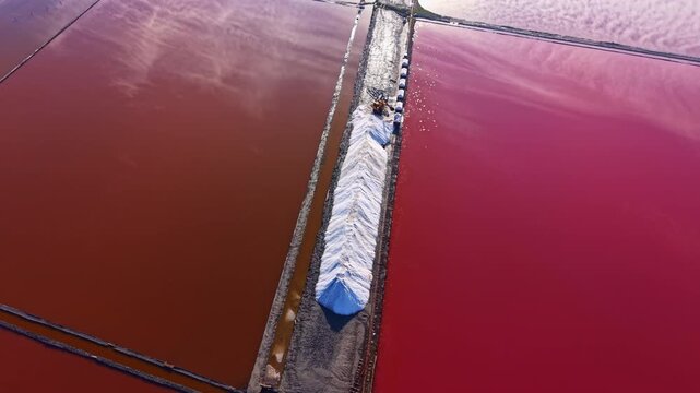 Colorful salt pans reflect brilliant shades of red and orange, forming a stunning contrast. Workers carry out the salt harvesting process against a backdrop of vibrant scenery.