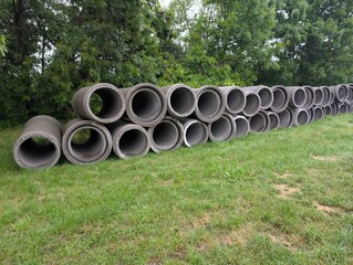 Rows of large, cylindrical concrete drainage pipes stacked on a grassy area near a forested background. Industrial materials in a natural environment. Contrast between infrastructure and greenery.