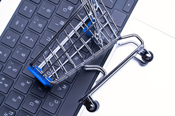 A shopping cart is on a laptop keyboard. The cart is made of metal and has a blue handle. The laptop is on a white surface
