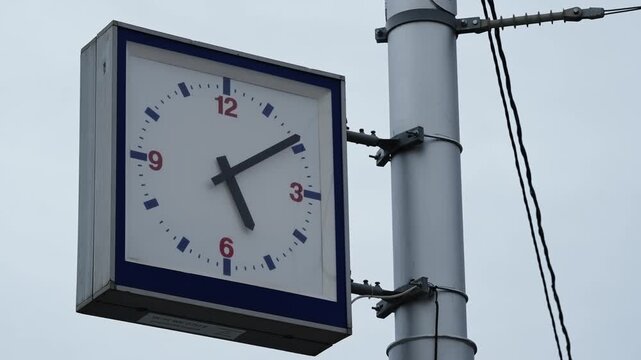 Close-up of a square outdoor analog clock mounted on a metal pole, red numerals and dark hands against a pale sky, with nearby utility cables, shown across two frames from a video