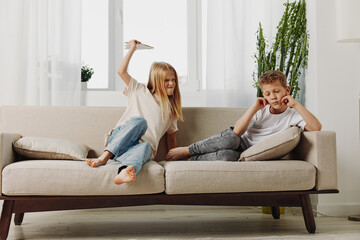 Obraz premium Two children sitting on beige sofa in bright living room, girl holding paper plane above boy while he covers ears. Concept of sibling interaction, playfulness, and childhood moments.
