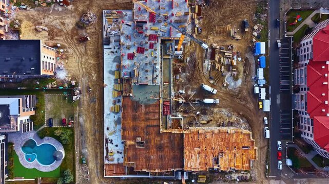 Workers and equipment are present at a construction site where a large building is being built. Cranes lift materials while trucks are parked nearby.
