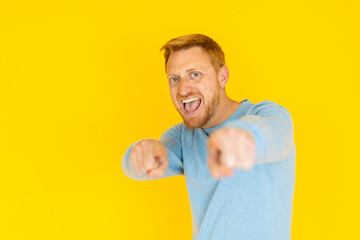 Excited redhead man pointing forward and shouting on a vibrant yellow background, expressing enthusiasm and confidence