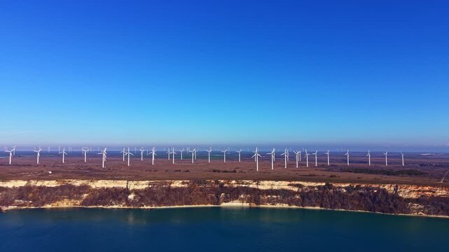 Wind turbines generate energy on land that meets the coastline, while clear blue skies and calm waters create a simple landscape view.