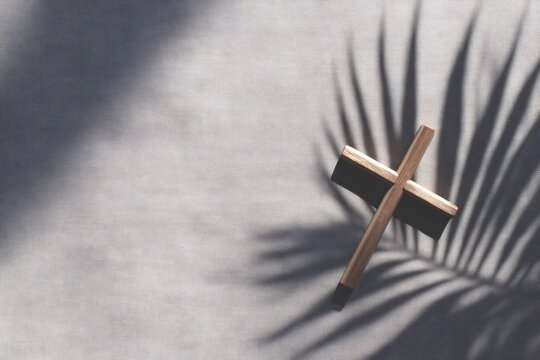religious still life, tidy ash wednesday flat lay small wood cross on gray linen, single palm frond shadow, soft morning light, large negative space above