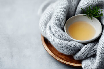 cozy winter still life, neatly displayed soft grey scarf on wooden tray, with pine sprig and small bowl of honey on a creamy surface, plenty of room for text