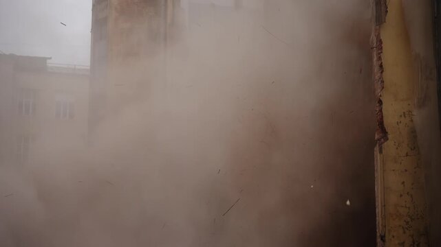 Old brick building undergoing demolition as exterior wall collapses, sending heavy dust and debris billowing, obscuring windows and facade in a dense cloud amid structural failure