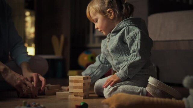 Young daughter and her mother playfully build fragile Jenga tower on floor, basking in warm evening atmosphere, promoting interactive learning and entertainment through this balancing board game fun