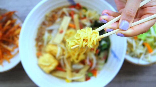 Woman's hand using chopsticks to pick up delicious yellow noodles from a steaming hot bowl of asian ramen soup with broth, tofu, and fresh vegetables in an authentic japanese restaurant