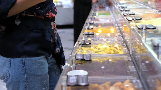 Close up of a female customer's hand selecting and taking a piece of dried mango from a transparent bulk bin dispenser in a grocery store, promoting healthy eating and zero waste lifestyle