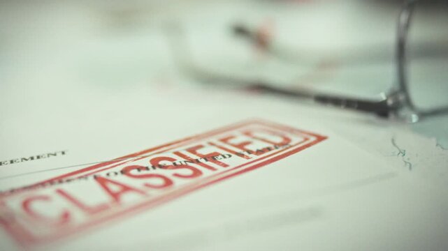 Classified documents bearing a prominent red confidentiality stamp are laid out on a table, visible alongside glasses and other classified papers in the background