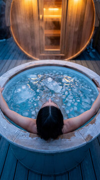 Woman enjoying an ice bath outdoors next to a warmly lit sauna, creating a serene spa-like environment focused on relaxation and wellness therapy. Image made using Generative AI.
