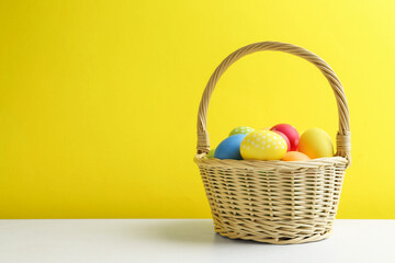 Painted Easter eggs in wicker basket on white table against yellow background. Space for text