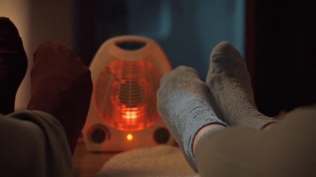 A cozy young couple in warm socks unwinds beside a radiant heater on a chilly evening, basking in the comforting warmth that emanates from the electric device