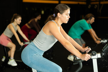 Naklejka premium Athletic woman in a top and leggings is pedaling an exercise bike in the gym. European woman doing a cardio circuit workout on the machine during a Crossfit workout