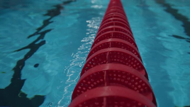 Close-up of a red swimming pool lane divider stretching into blue water, with droplets on the surface and blurred swimmers in the background, capturing motion, focus, and a competitive training