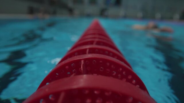 Close-up of a red swimming pool lane divider stretching into blue water, with droplets on the surface and blurred swimmers in the background, capturing motion, focus, and a competitive training