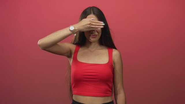Young hispanic woman covering eyes with hand in pink studio setting; shyness privacy vulnerability intimacy.