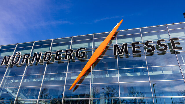 Au&szlig;enansicht des gl&auml;sernen Eingangsbereichs der N&uuml;rnberg Messe mit dem markanten orangefarbenen Logoelement an der Fassade. 
