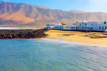 Coastal village of Famara, Lanzarote, with white houses and green shutters facing a golden beach....