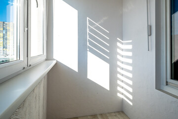 Obraz premium Interior of a modern balcony in light colors. Sunlight casts geometric shadows through the blinds onto the white wall.