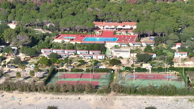 SAN VITO LO CAPO, SICILY, ITALY, 24 JAN 2026: AERIAL: Seaside campground resort with large swimming pool among dense pine trees, with rows of cabins and areas for sports activities near rocky beach.