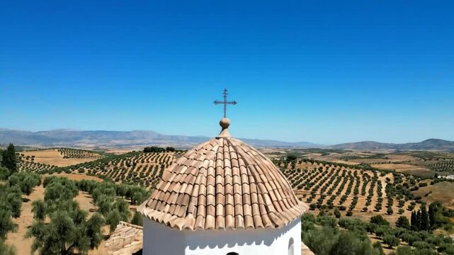 Scenic white chapel with a cross on top surrounded by rows of olive trees and mountains under a blue sky
