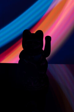 Silhouette of Maneki-neko Lucky Cat against Vibrant Light Painting Background, Mysterious Dark Fortune Cat Figurine with Colorful Cyberpunk Light Trails