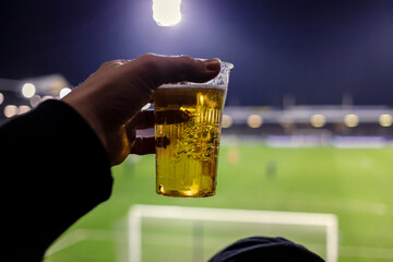 Hand holding plastic cup of beer at football stadium with blurred pitch and floodlights in background, fan perspective © Victor