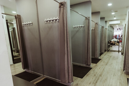 Interior of a clothing store fitting room featuring multiple gray changing stalls with curtains and wooden flooring, illuminated by bright overhead lights