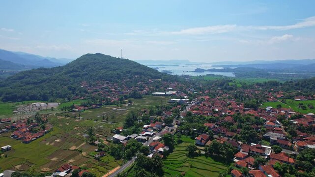 Aerial drone footage of a local village with main roads, a hill, with forests and flat wide rice fields, and the Jatigede reservoir behind, in Wado area, Sumedang regency, Java island, Indonesia