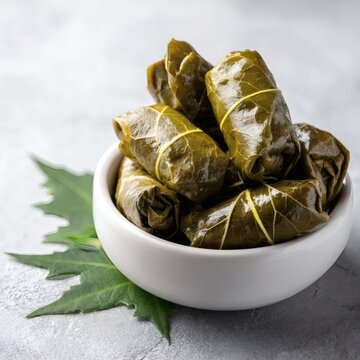 Delicious dolmas wrapped in vine leaves, tied with threads, placed in a white ceramic bowl, on a light gray textured surface, with a few fresh leaves beside, in a traditional Mediterranean c