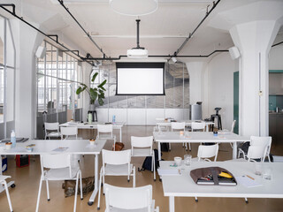Fototapeta premium Modern meeting room with projector screen, white tables and chairs, industrial ceiling and plants, training space