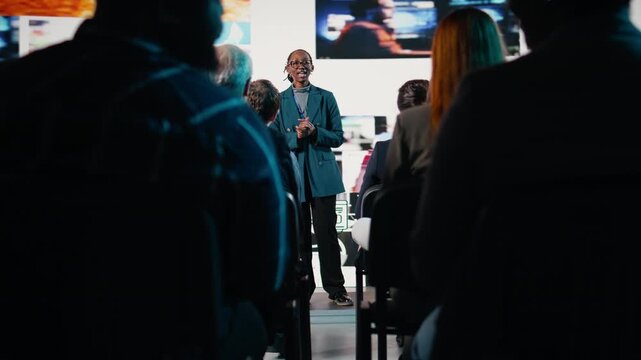 African American female speaker delivers powerful media presentation at tech conference, diverse audience in modern conference hall. Young adult discusses advertising and public relations. Camera A.