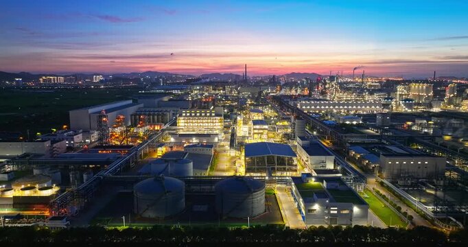 Aerial shot of a large industrial chemical plant and refinery complex glowing with lights in an urban industrial zone during a beautiful sunset at dusk.