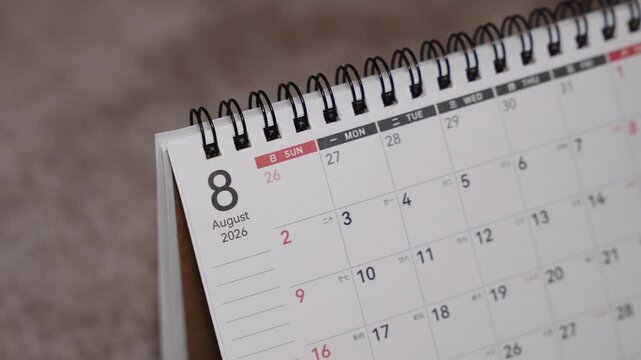 Close-up of a desk calendar page being flipped from July 2026 to August 2026. Hand turns the paper forward while the camera remains static, presenting a clear summer month time transition indoors.