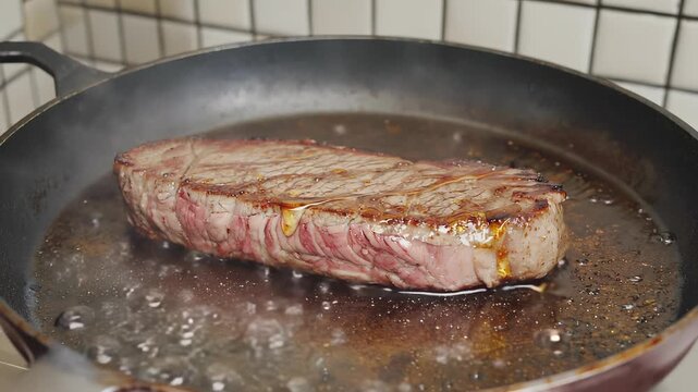Steak fat edge cooks slowly in a hot pan. Golden droplets form and crackle as steam gently rises around. The tiled backsplash adds to the kitchen atmosphere while the heat glows beneath