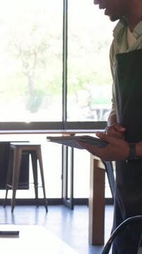 Vertical video: Entering right server in apron tapping tablet and phone beside window table selling