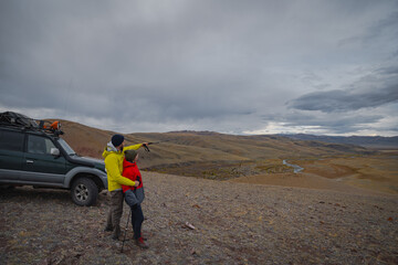 Two hikers stand by an off-road vehicle, hugging and pointing at a rugged mountain landscape under a cloudy sky during their autumn adventure.