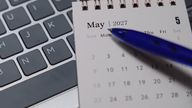 Close-up of a desk calendar on a laptop keyboard as a blue pen points to May 2027. Static camera presents focused date indication and late spring monthly planning concept in a modern office setting.