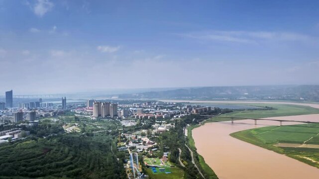 Aerial time-lapse of Sanmenxia cityscape alongside the Yellow River, featuring modern buildings, green parks, bridges, and winding river under a clear sky, Henan Province, China.