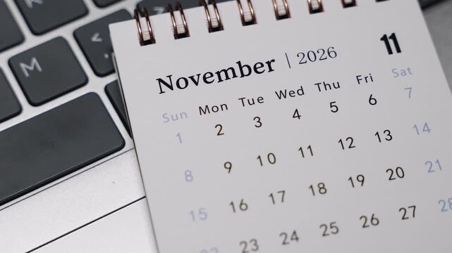 Close-up of a desk calendar displaying November 2026 on a laptop keyboard. Camera slowly rotates to the right, emphasizing monthly dates and structured time planning in a professional office environm