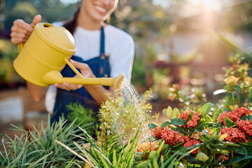 Woman wearing an apron holding a yellow watering can, irrigating plants and flowers in a outdoor plant nursery © we.bond.creations