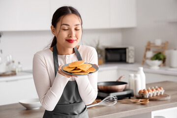 Beautiful young happy Asian woman with plate of tasty pancakes in kitchen