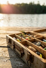 Fototapeta premium Rustic fishing tackle box with a fly lure on a wooden dock by a serene lake at golden hour, evoking peaceful outdoor recreation.