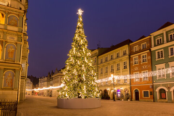 Fototapeta premium Large decorated tree shines in the center of Poznan s historic square, surrounded by colorful buildings and festive lights.