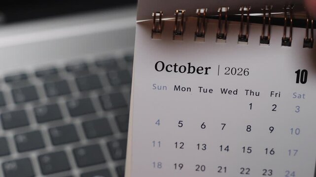 Close-up of a desk calendar on a laptop keyboard as a hand flips the page from September 2026 to October 2026. Camera stays static, presenting a clear monthly time transition and organized planning c